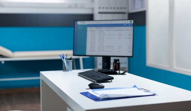 Text: Empty doctor's office desk with computer and medical exam bed 